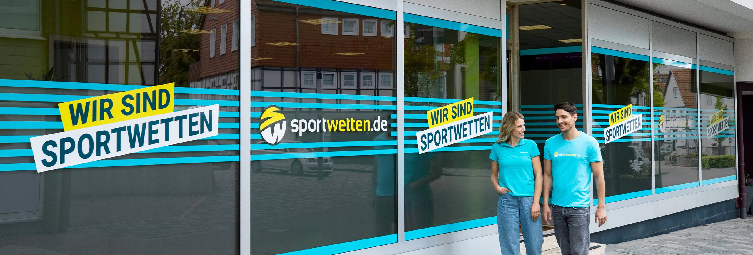 Ein Mitarbeiter und eine Mitarbeiterin in türkisfarbenen T-Shirts, mit sportwetten.de-Logo,  
laufen vor einem sportwetten.de-Wettbüro mit gläsernen Schaufenstern vorbei. Auf dem Schaufenstern  
befindet sich ein Schriftzug „Wir sind sportwetten“ und das Logo von sportwetten.de.